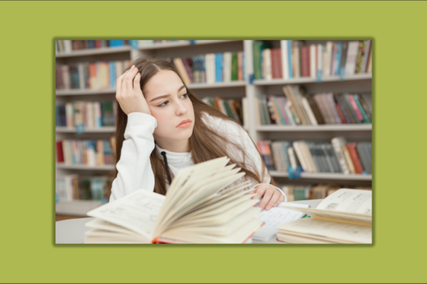 young girl at a table in a library with books open, leaning on her hand, looking off into the distance. 