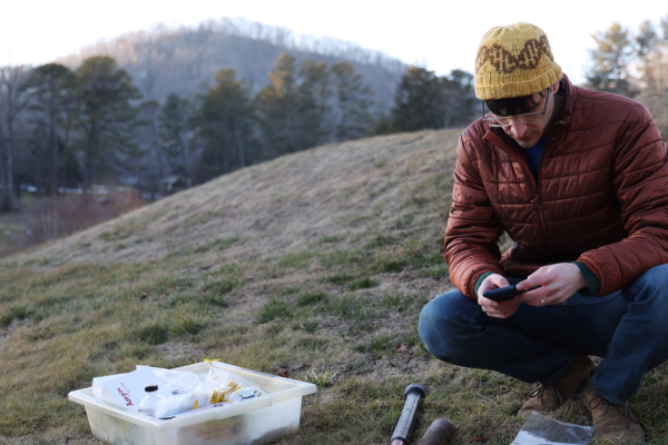 Julian Liber, a fourth-year PhD student in hat and puffy jacket, sitting on the side of a mountain collecting fungal samples. 