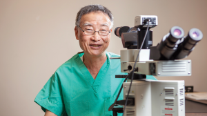 Tai-Po Tschang, MD’72 in green scrubs sitting at a large complex microscope