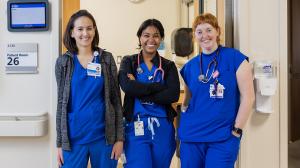 Three female health providers (young, 20s or 30s) in Duke blue scrubs stand in a patient room doorway. They smile at the camera. 