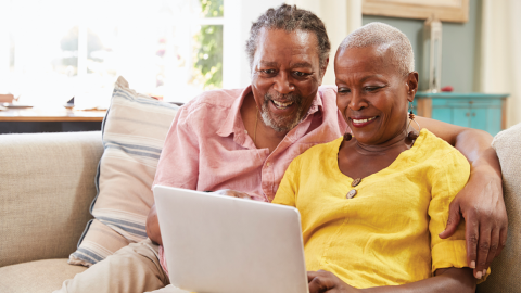 an older Black couple sitting together on a sofa sharing a laptop. 